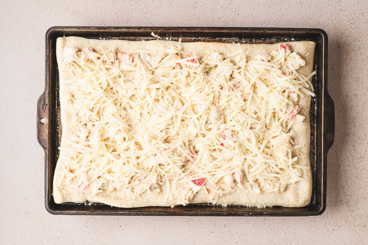 Unbaked crab rangoon pizza on a sheet pan topped with creamy crab mixture and shredded mozzarella.
