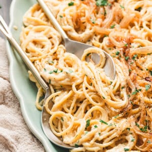 Close-up of creamy caramelized onion pasta on a green serving platter, garnished with fresh parsley and black pepper, with a spoon and fork ready for serving.