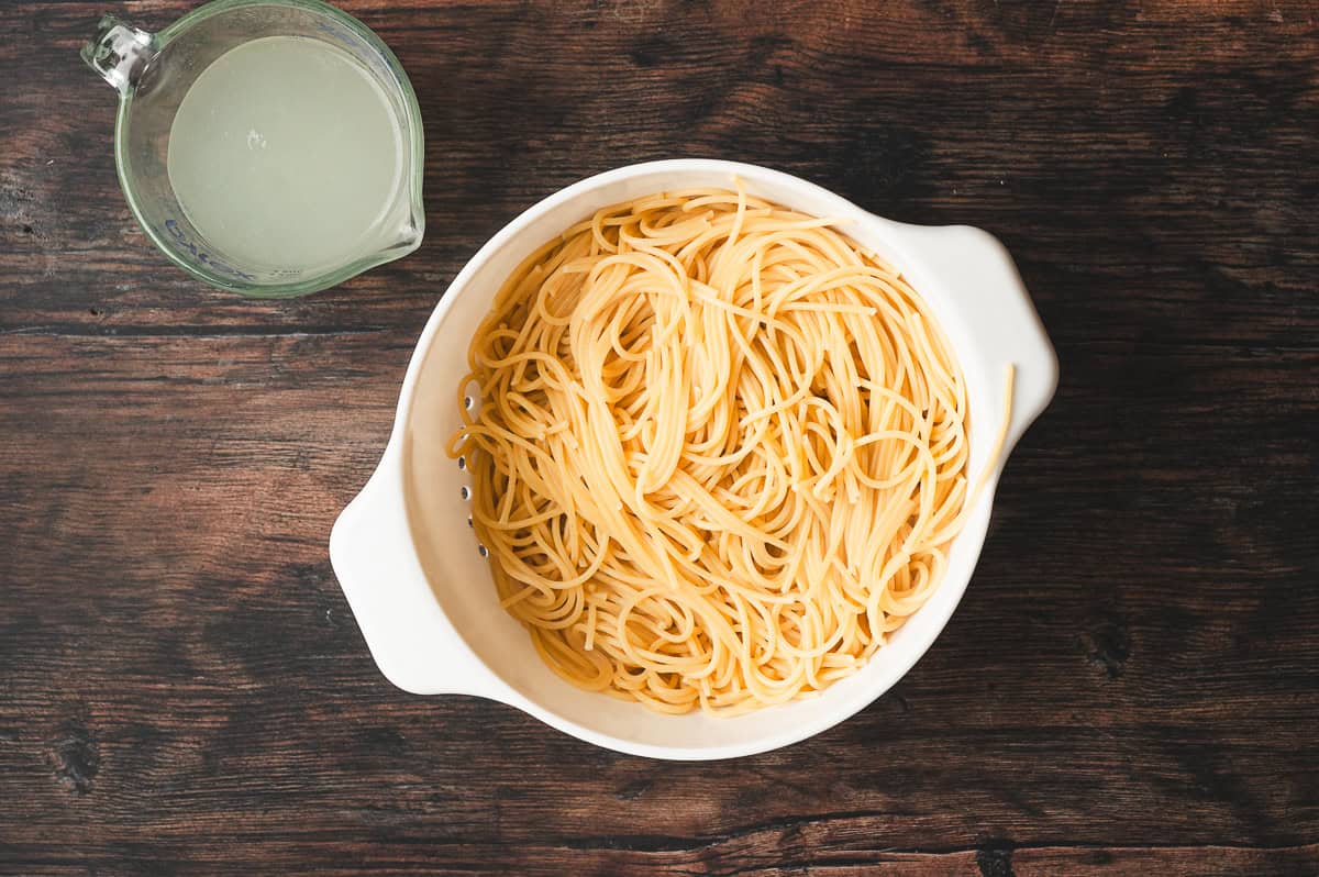 Cooked spaghetti drained in a white colander with a measuring cup of reserved starchy pasta water on the side.