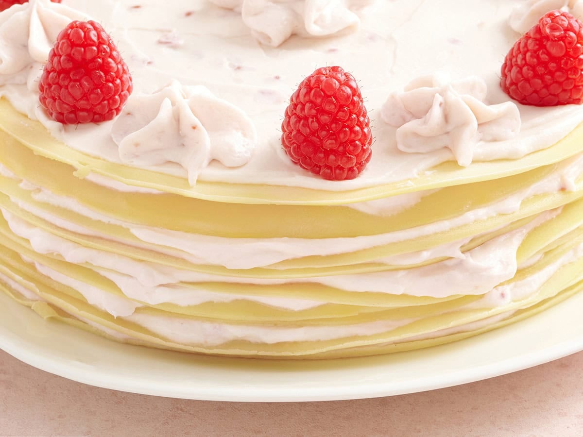 Close-up of the side of a crepe cake, showing thin crepe layers and raspberry cream filling, topped with piped cream swirls and raspberries.