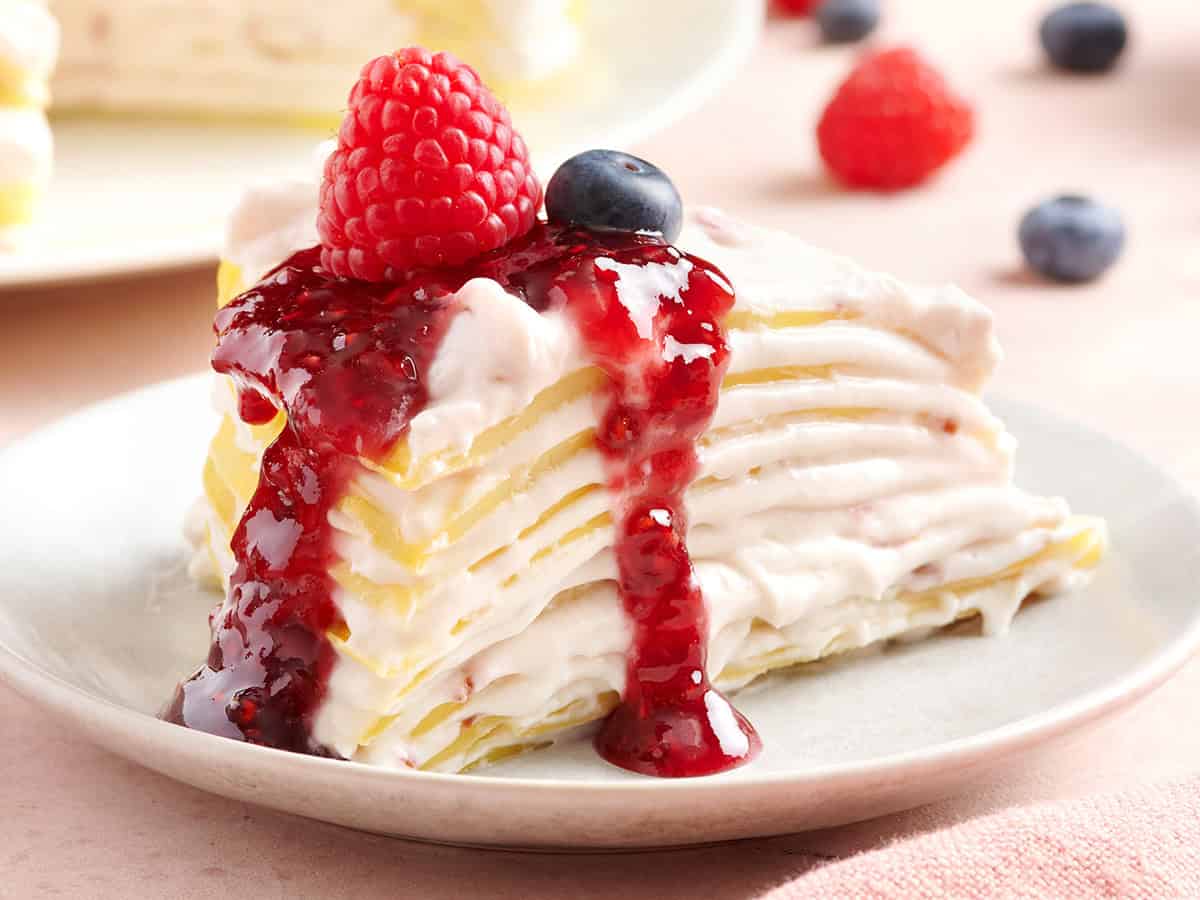 A slice of crepe cake topped with raspberry jam sauce, fresh raspberry, and blueberry on a white plate, with more berries scattered in the background.