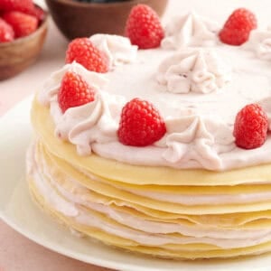 A layered crepe cake filled with raspberry cream, topped with piped cream swirls and fresh raspberries, with bowls of blueberries and raspberries in the background.