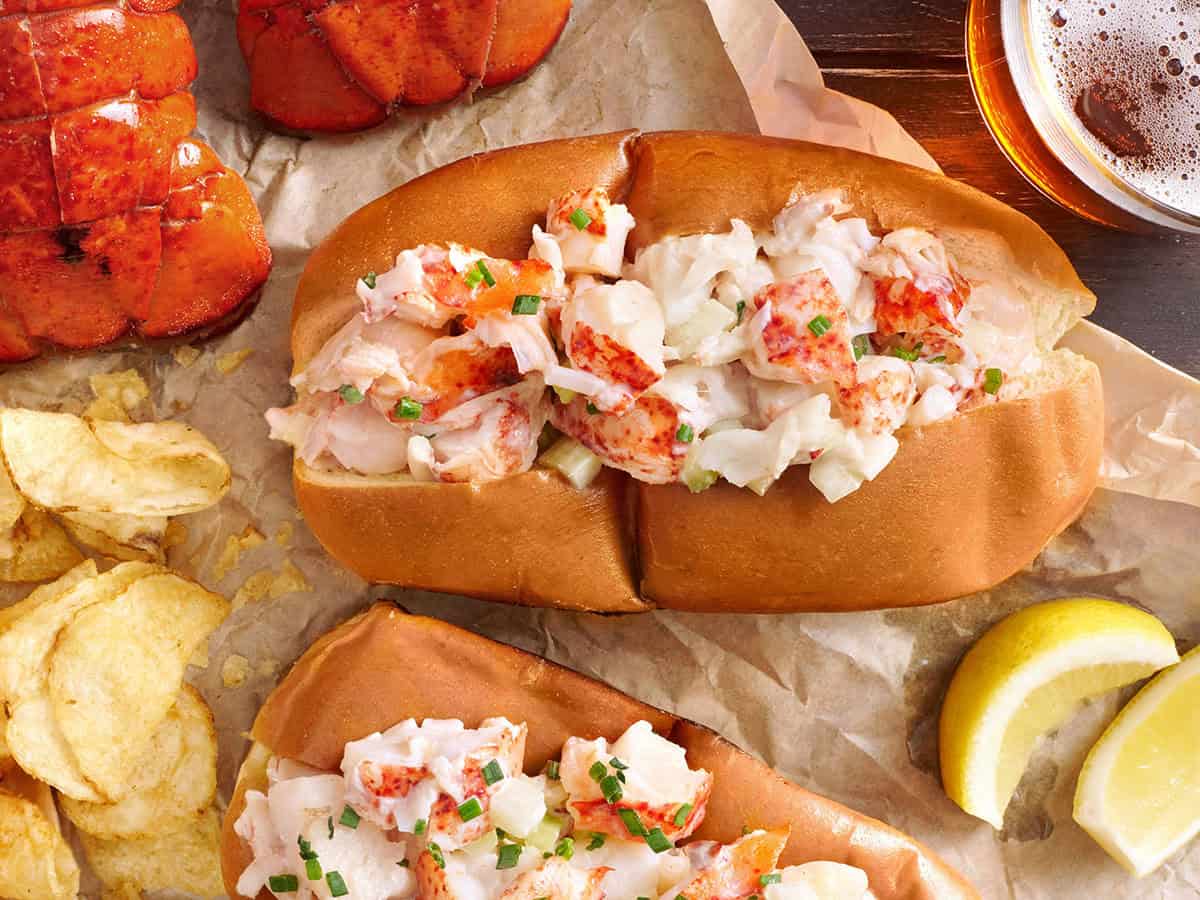 Lobster rolls on parchment paper served with crinkle fries, lemon wedges, and beer.