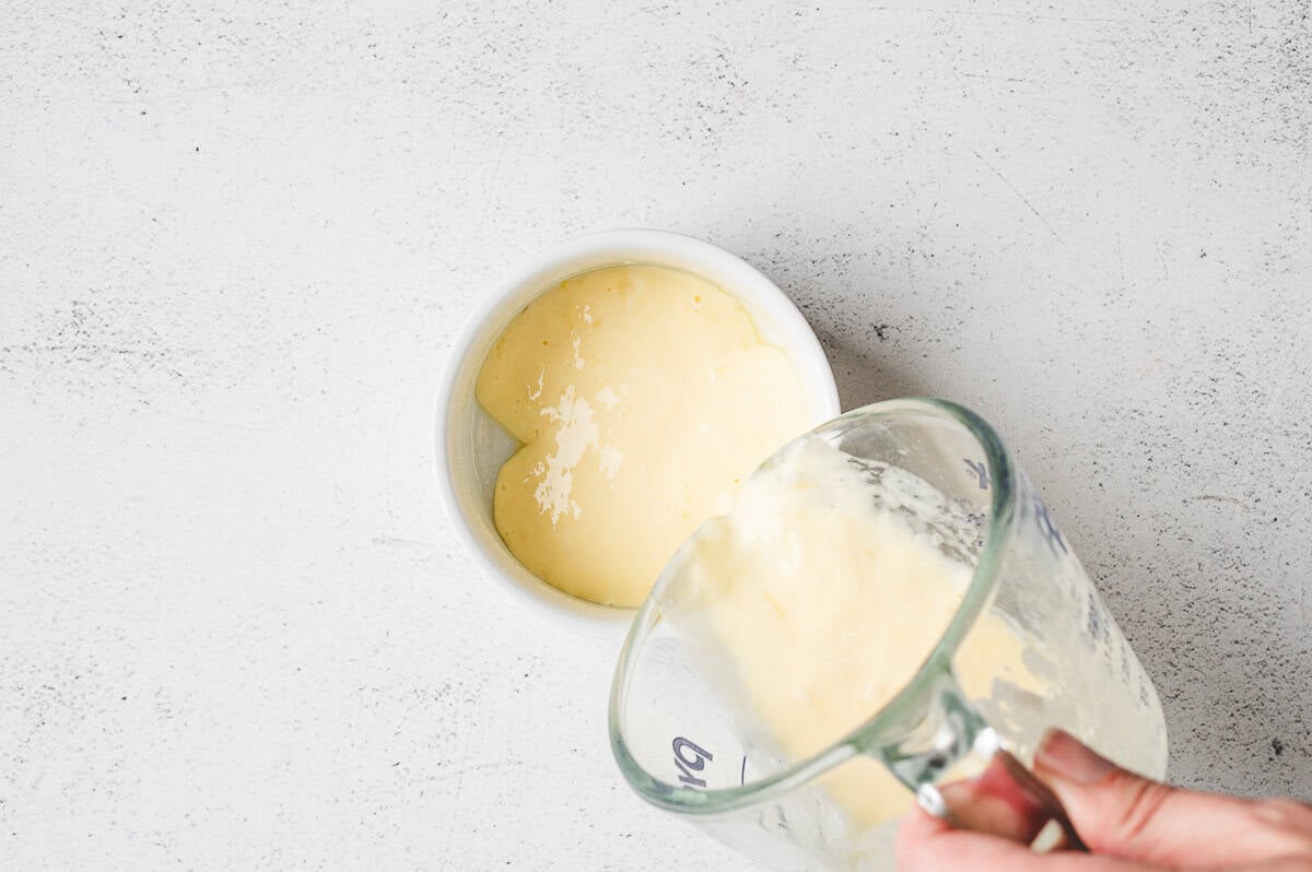 Hand pouring the whisked egg and mayonnaise mixture from a measuring cup into a greased white ramekin.