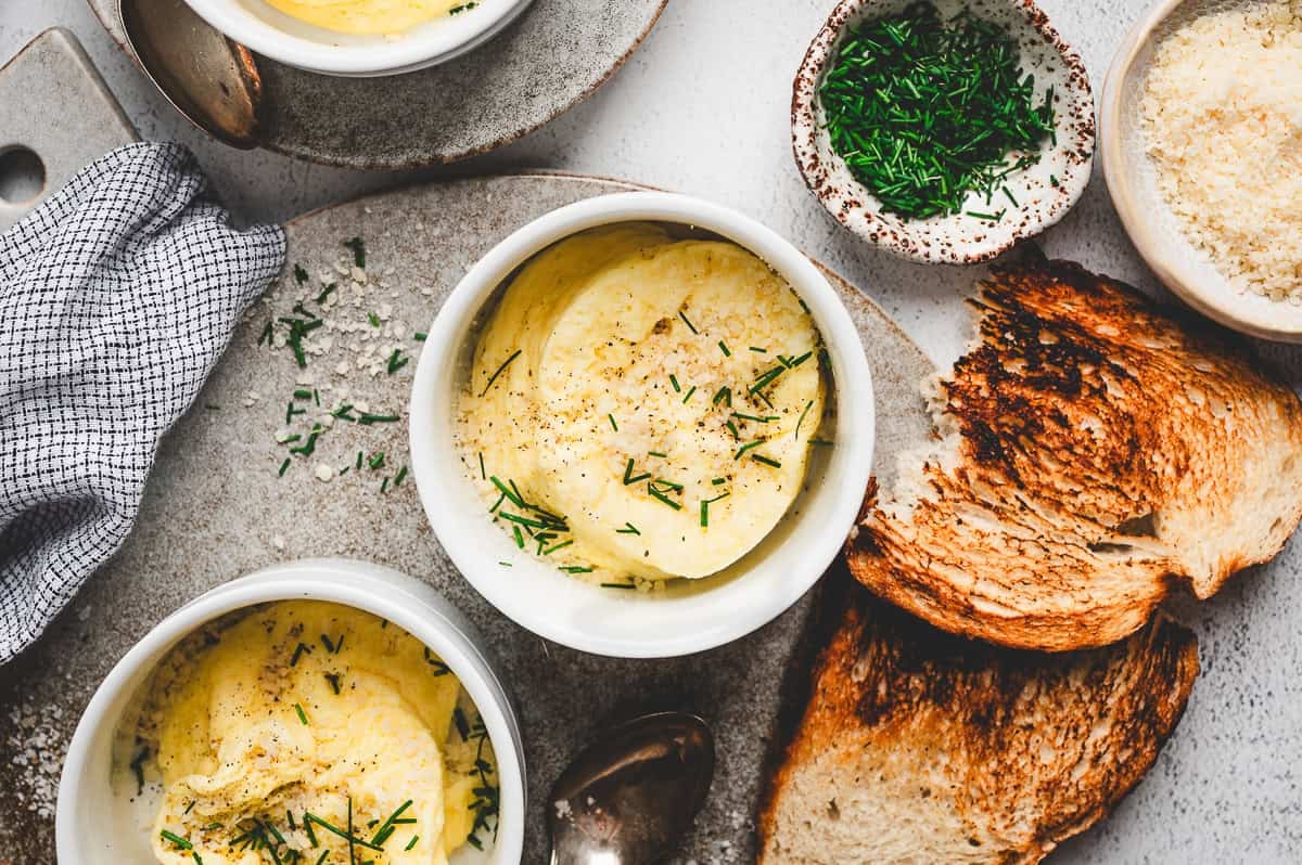 Overhead view of microwave soufflé-style eggs in ramekins, topped with Parmesan and chives, served with toasted bread on the side.