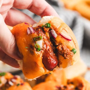 Close-up of a hand holding a mini chili cheese dog with melted cheddar, chili, diced red onion, and scallions on a Hawaiian roll.