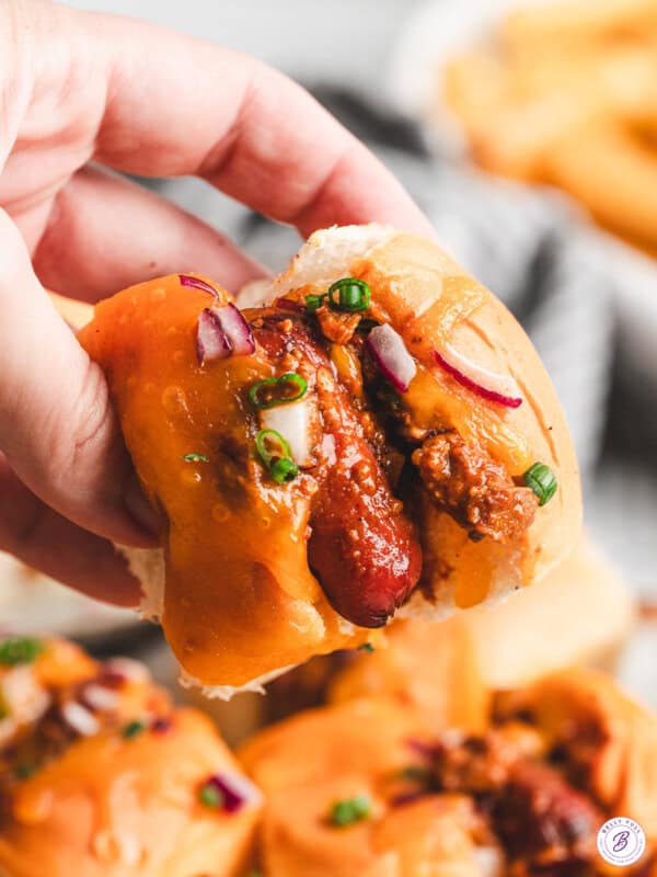 Close-up of a hand holding a mini chili cheese dog with melted cheddar, chili, diced red onion, and scallions on a Hawaiian roll.