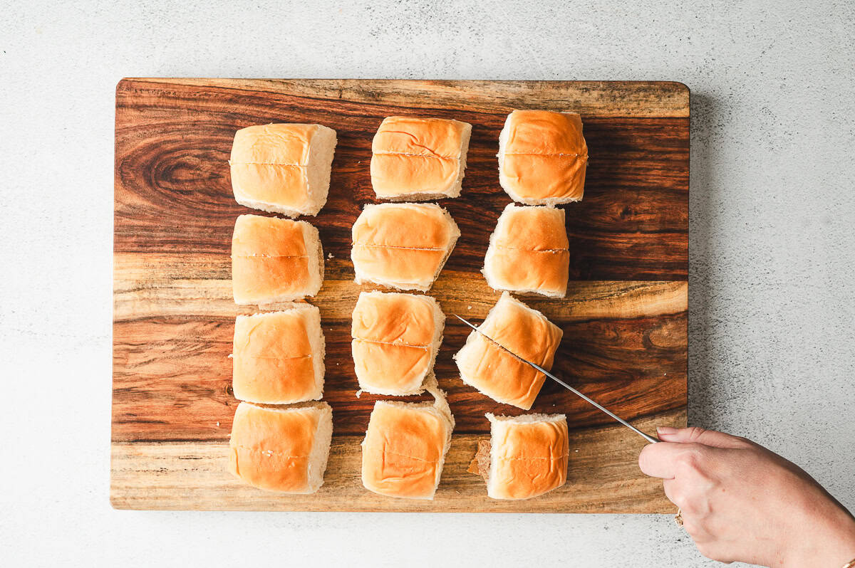 Sweet Hawaiian rolls being separated and sliced down the center on a wooden cutting board with a knife.