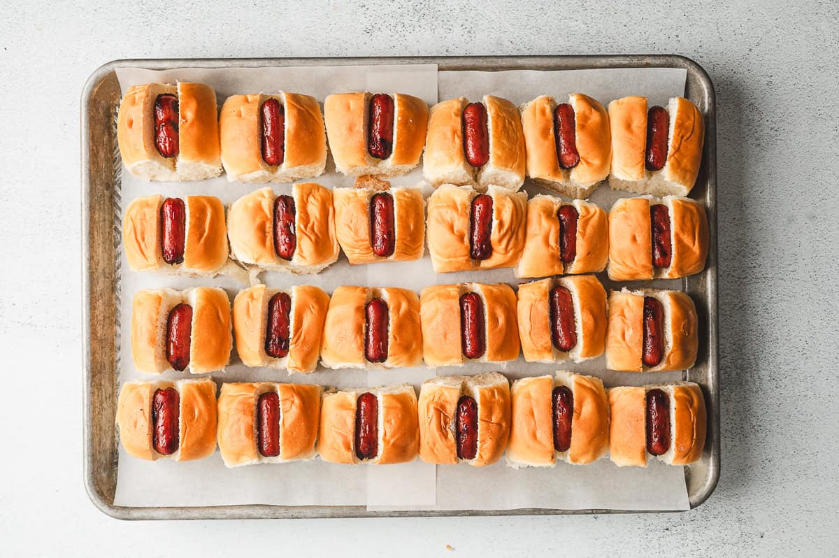 Cocktail franks tucked into sliced Hawaiian rolls, arranged neatly on a parchment-lined baking sheet.