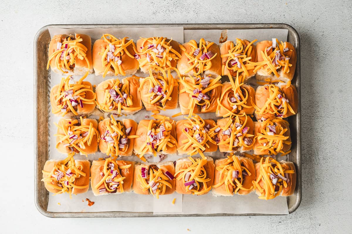Mini chili dogs topped with shredded cheddar cheese and finely diced red onion, ready to go into the oven.