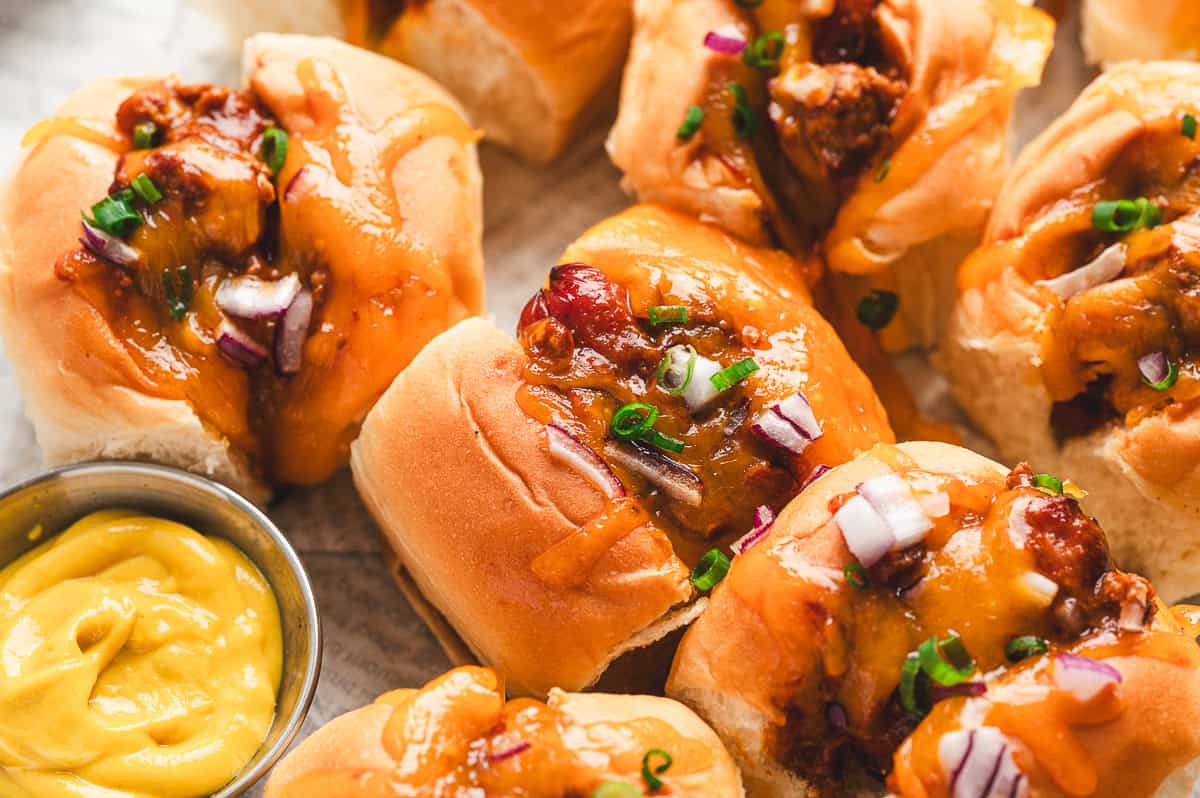 Golden mini chili cheese dogs on a tray, topped with melted cheese, red onion, and scallions, served with mustard on the side.