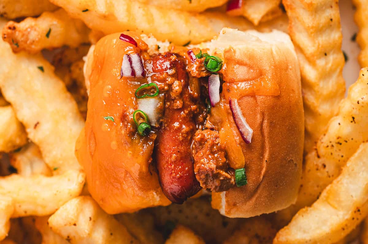 Mini chili cheese dog on a plate surrounded by seasoned crinkle-cut fries, topped with chili, cheddar, onions, and scallions.