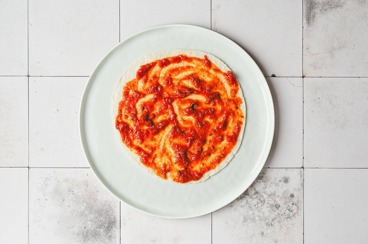 Flour tortilla on a plate spread with marinara sauce in an even layer.