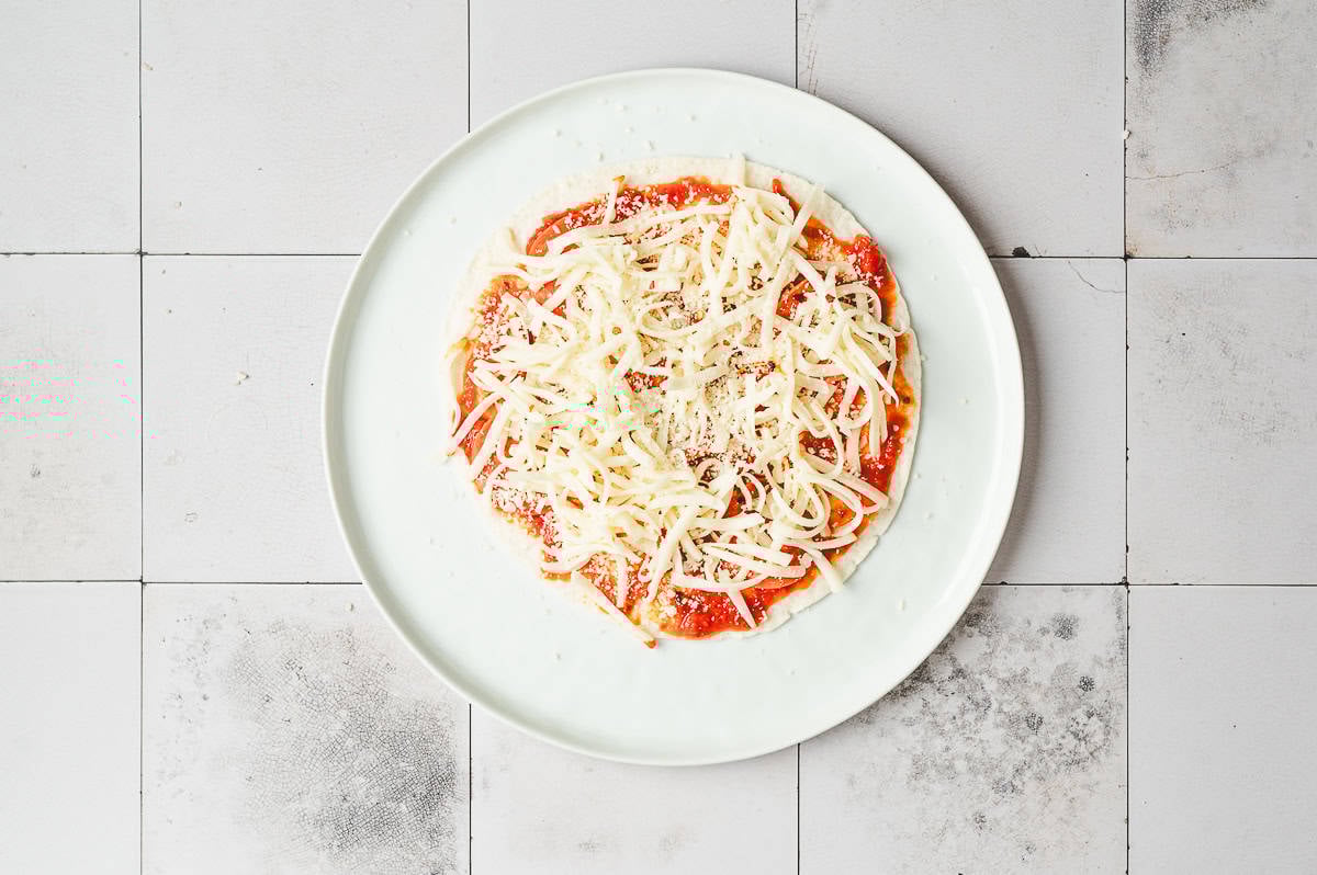 Tortilla topped with marinara sauce, shredded mozzarella, and Parmesan on a plate.