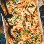 A baked Thai dumpling casserole in a white dish, filled with ramen noodles, dumplings, and a creamy curry sauce, garnished with scallions, cilantro, chili crisp, and sesame seeds.