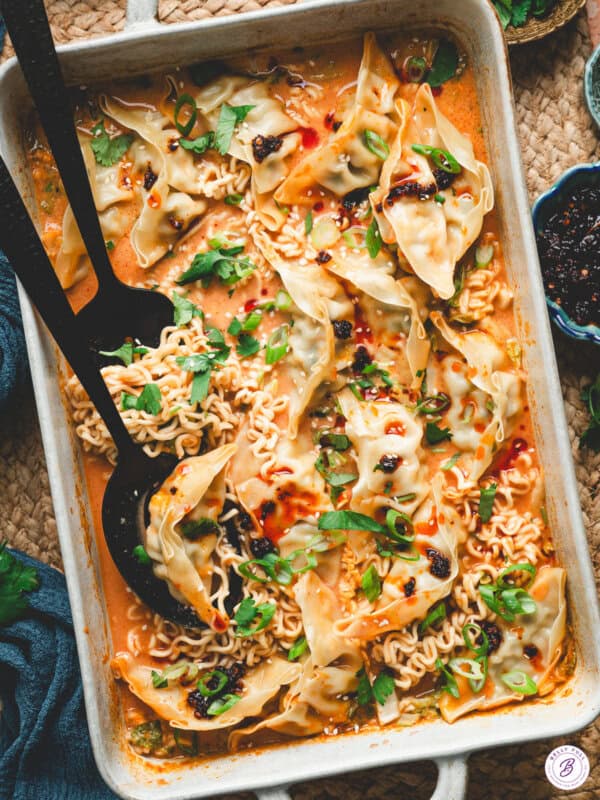 A baked Thai dumpling casserole in a white dish, filled with ramen noodles, dumplings, and a creamy curry sauce, garnished with scallions, cilantro, chili crisp, and sesame seeds.