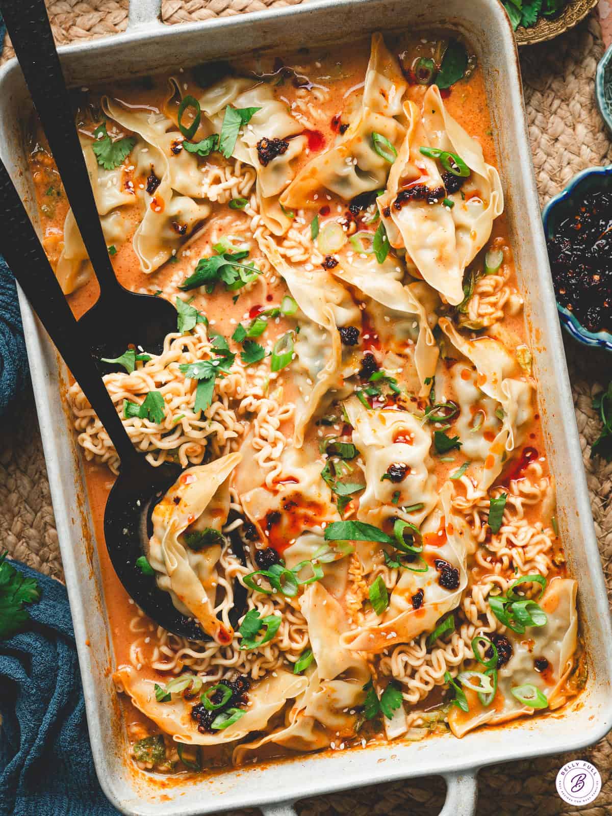 A baked Thai dumpling casserole in a white dish, filled with ramen noodles, dumplings, and a creamy curry sauce, garnished with scallions, cilantro, chili crisp, and sesame seeds.
