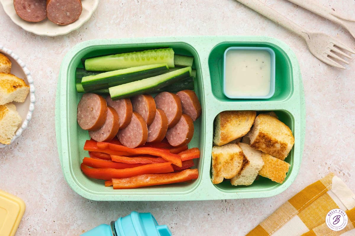 bento box for lunch with bread cubes, red bell pepper sticks, cucumber slices, sliced kielbasa, with Ranch dressing