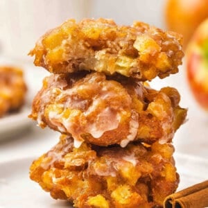 A stack of three glazed apple fritters on a white plate, the top one with a bite taken out, showing chunks of apple inside.