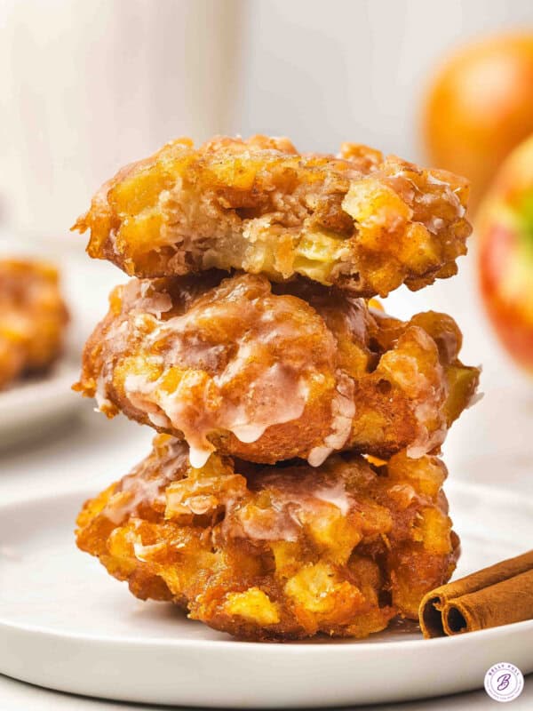 A stack of three glazed apple fritters on a white plate, the top one with a bite taken out, showing chunks of apple inside.