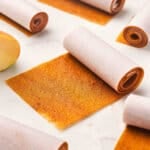 Rolled strips of homemade apple fruit leather wrapped in parchment paper, with one unrolled piece showing the chewy texture. Step 1: Bowl filled with peeled and chopped apples ready for cooking.