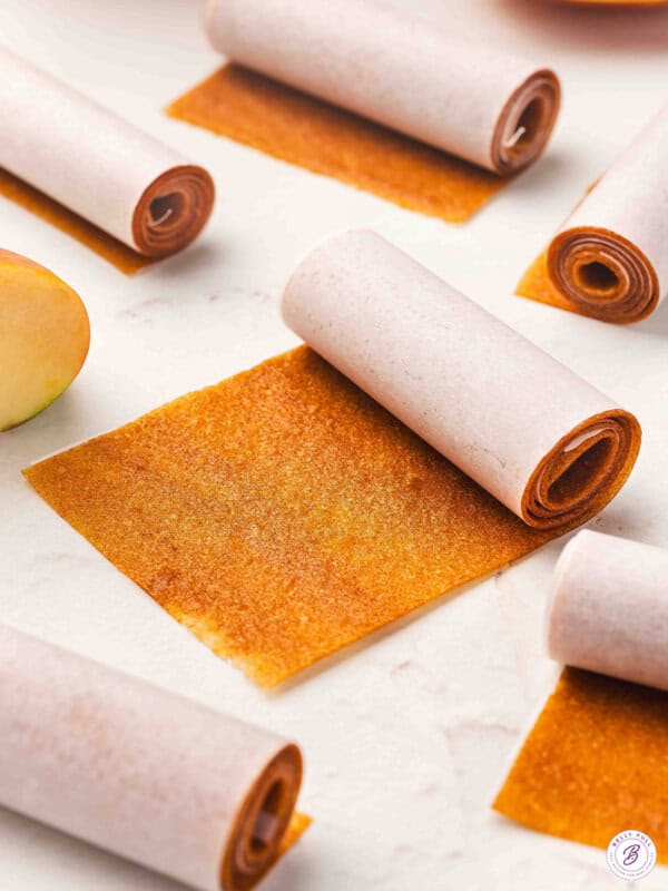 Rolled strips of homemade apple fruit leather wrapped in parchment paper, with one unrolled piece showing the chewy texture. Step 1: Bowl filled with peeled and chopped apples ready for cooking.