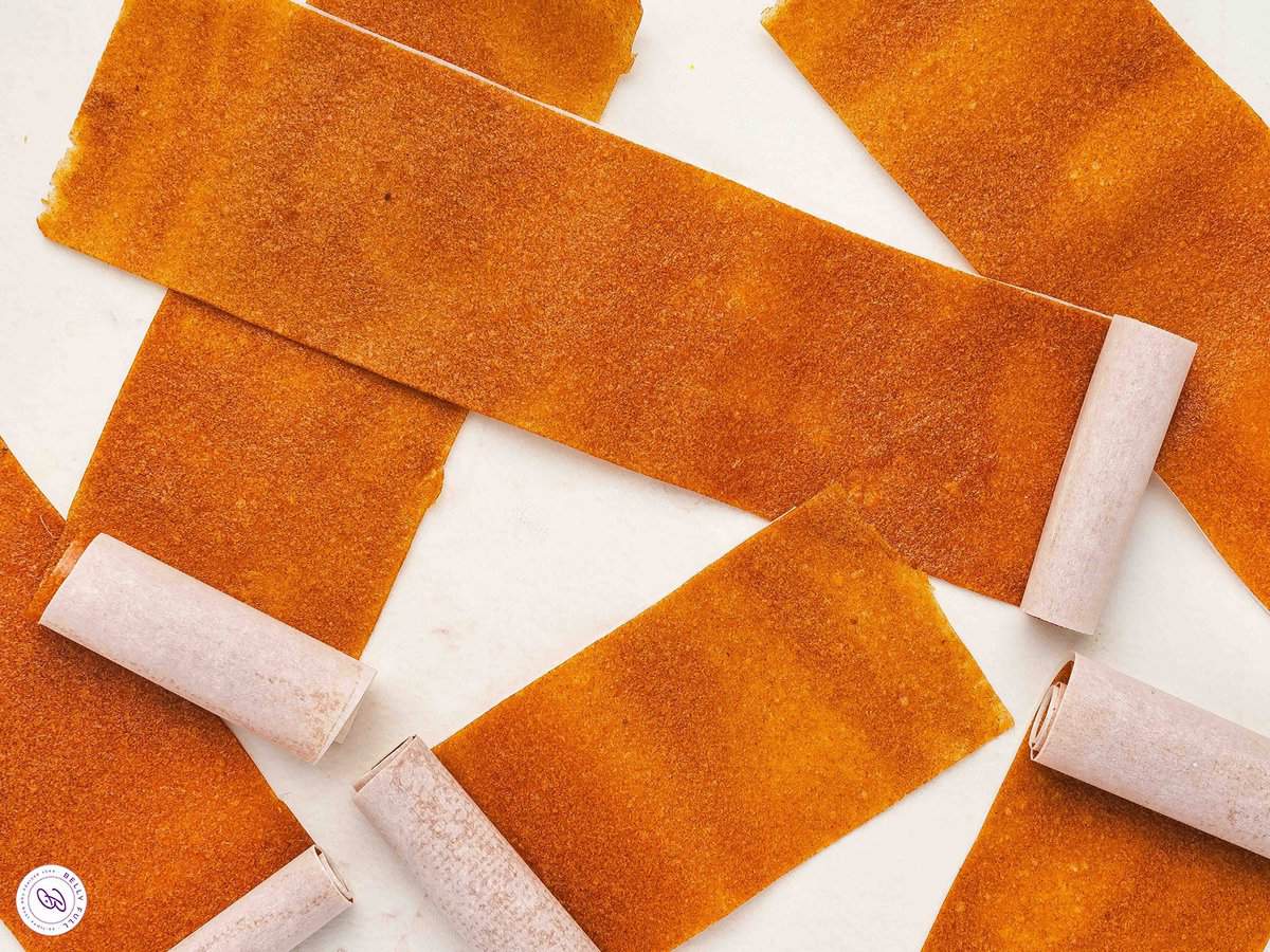 Sheets and rolls of apple fruit leather arranged on a white surface, showing the chewy texture and deep golden-orange color.