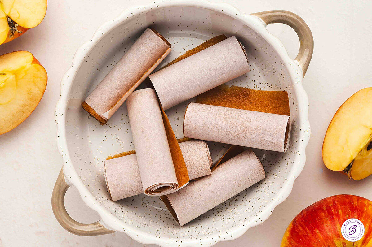 Rolled strips of homemade apple fruit leather wrapped in parchment paper, placed in a white speckled bowl with fresh apple halves around it.