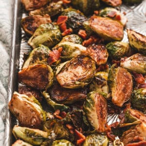 Roasted brussels sprouts with crispy edges, tossed with balsamic vinegar and crumbled bacon, served on a silver baking sheet.