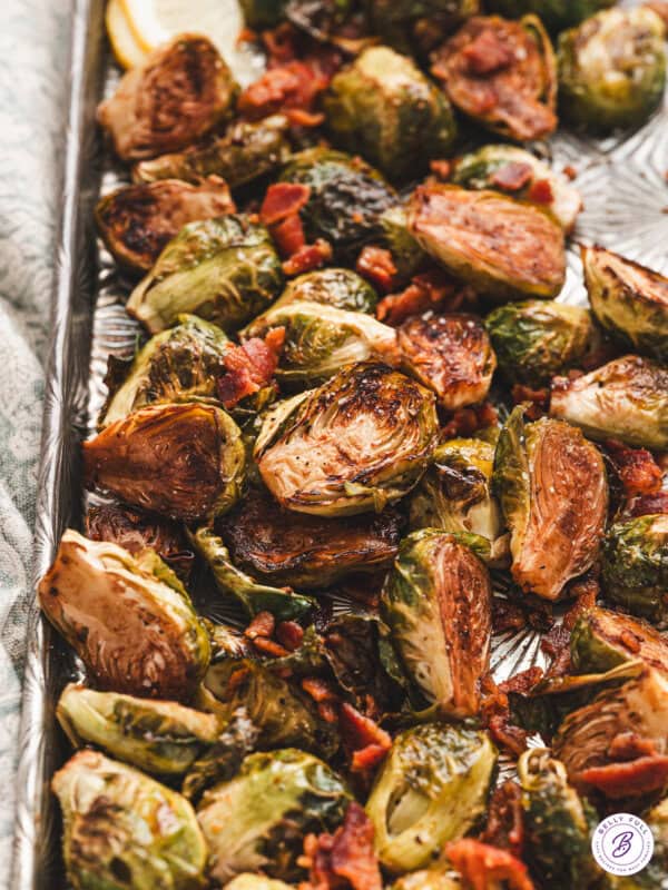 Roasted brussels sprouts with crispy edges, tossed with balsamic vinegar and crumbled bacon, served on a silver baking sheet.