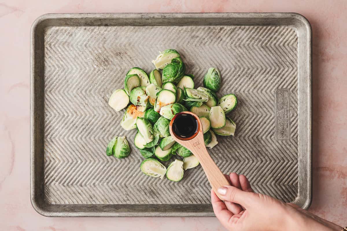 Halved raw brussels sprouts on a baking sheet with a hand holding a wooden tablespoon filled with balsamic vinegar, ready to drizzle.