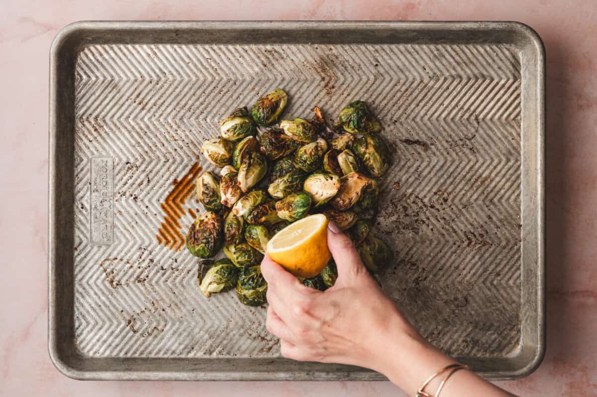 A hand squeezing half a lemon over roasted brussels sprouts on a baking sheet.