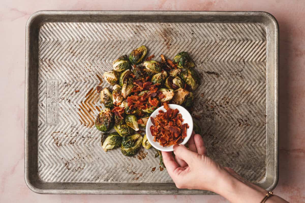 A hand adding a small white bowl of crumbled crispy bacon to roasted brussels sprouts on a baking sheet.