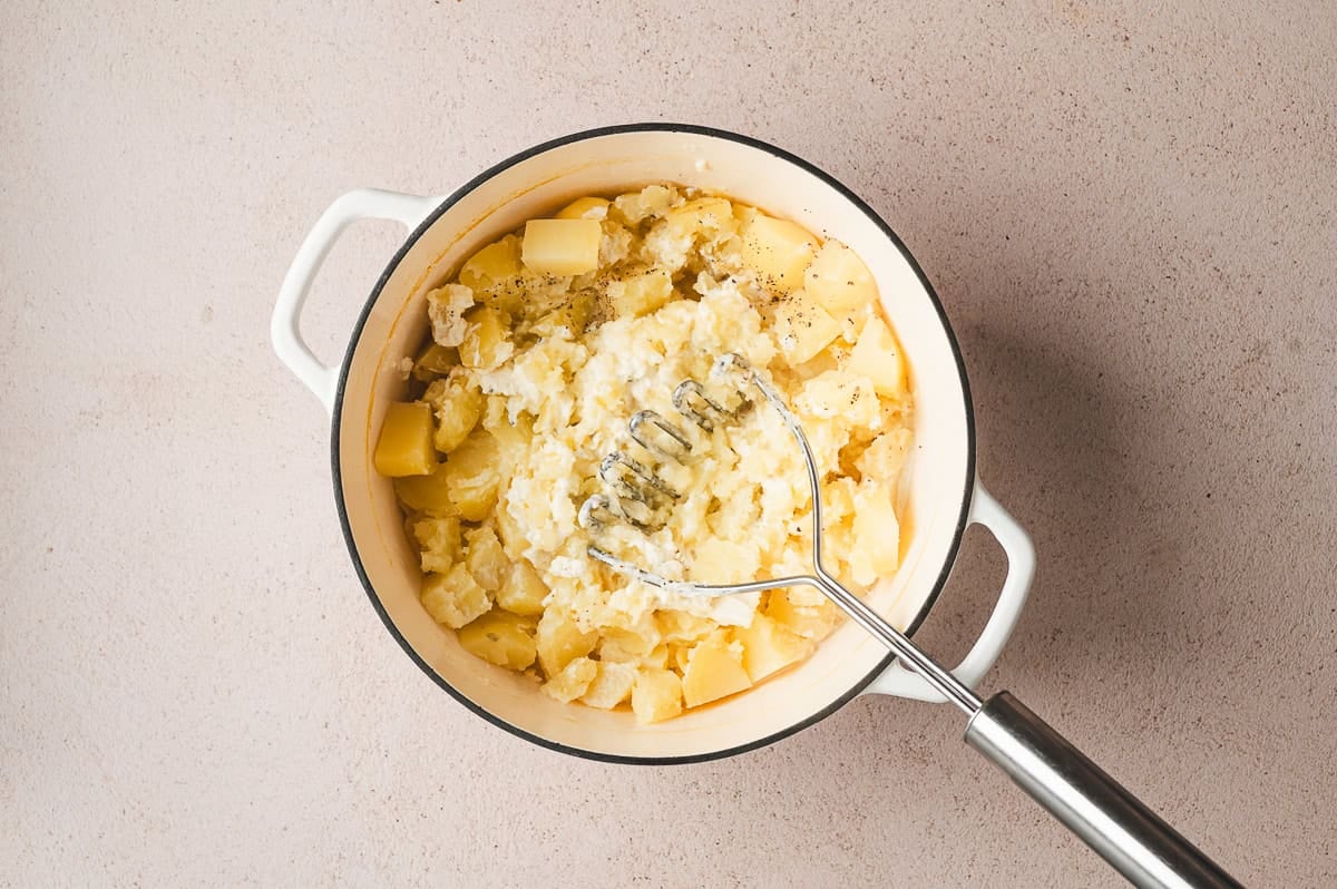Potatoes in a Dutch oven with Boursin cheese and pepper, ready to be mashed with a potato masher.