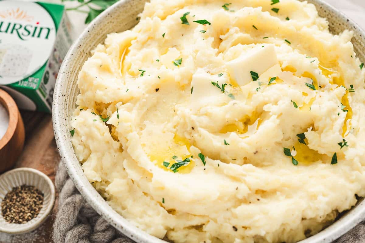 Close-up of mashed potatoes in a serving bowl with pats of butter, parsley garnish, and a box of Boursin cheese visible in the background.