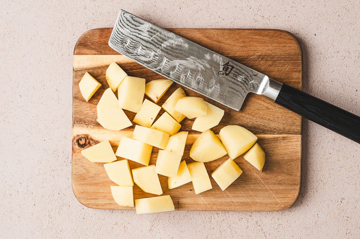 Peeled Yukon Gold potatoes cut into chunks on a wooden cutting board with a chef’s knife.