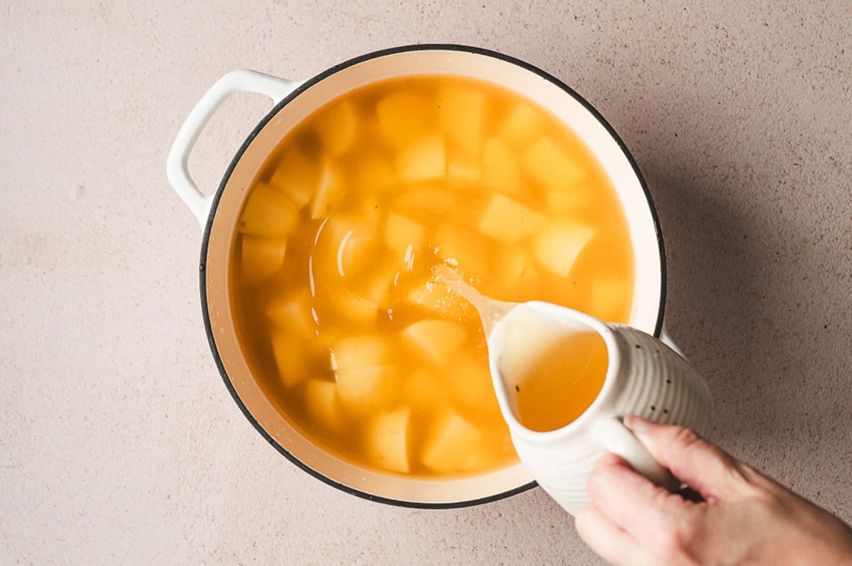 Potato chunks in a large pot being covered with chicken broth from a white pitcher.