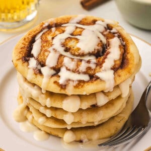 A tall stack of cinnamon roll pancakes drizzled with cream cheese icing on a white plate with a fork, with glasses of juice and cinnamon sticks in the background.