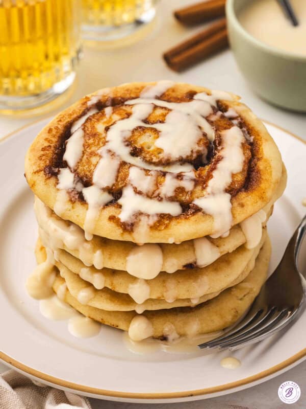 A tall stack of cinnamon roll pancakes drizzled with cream cheese icing on a white plate with a fork, with glasses of juice and cinnamon sticks in the background.