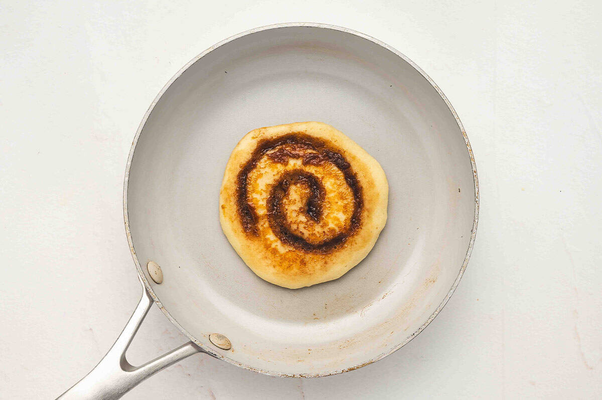 A golden-brown pancake in a skillet showing the cooked cinnamon swirl baked into the surface.