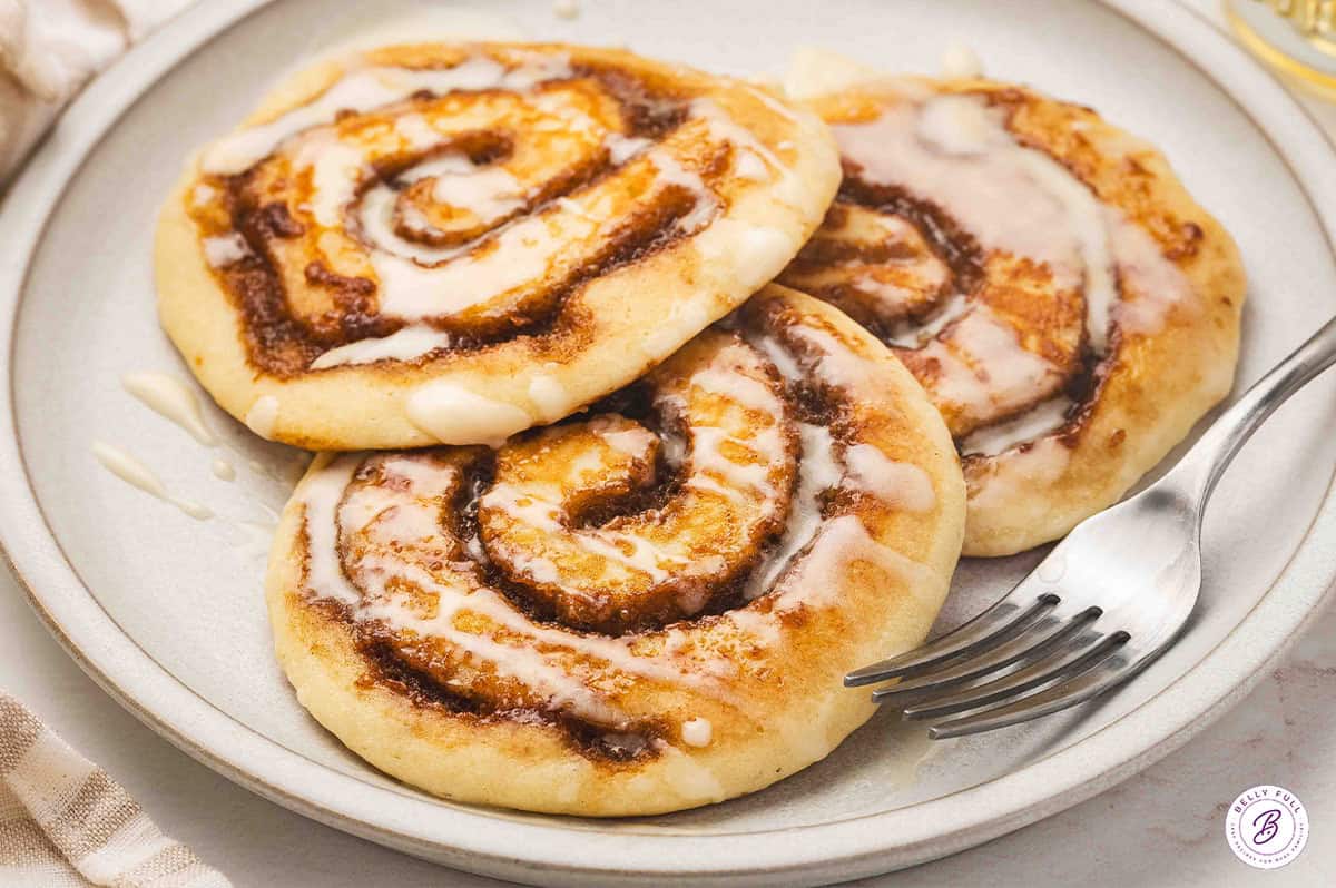 Three cinnamon roll pancakes topped with cream cheese icing on a plate with a fork, ready to serve.