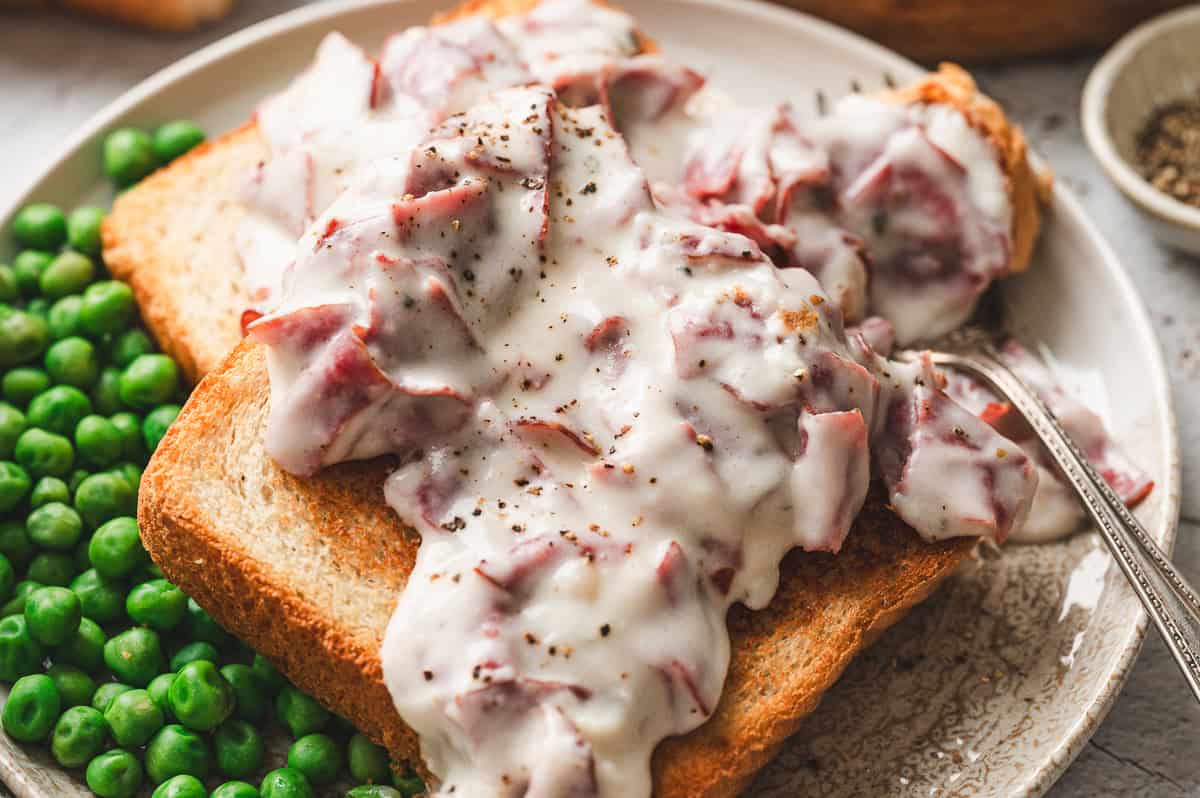 A plate of toast topped with creamed chipped beef, served alongside bright green peas with a fork on the side.