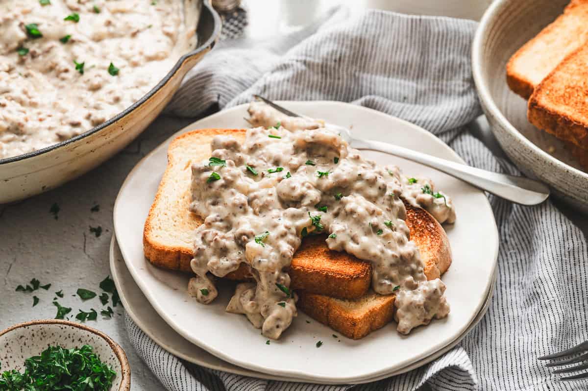 Plate of toasted white bread topped with generous scoops of creamy hamburger gravy, served with a fork on the side.
