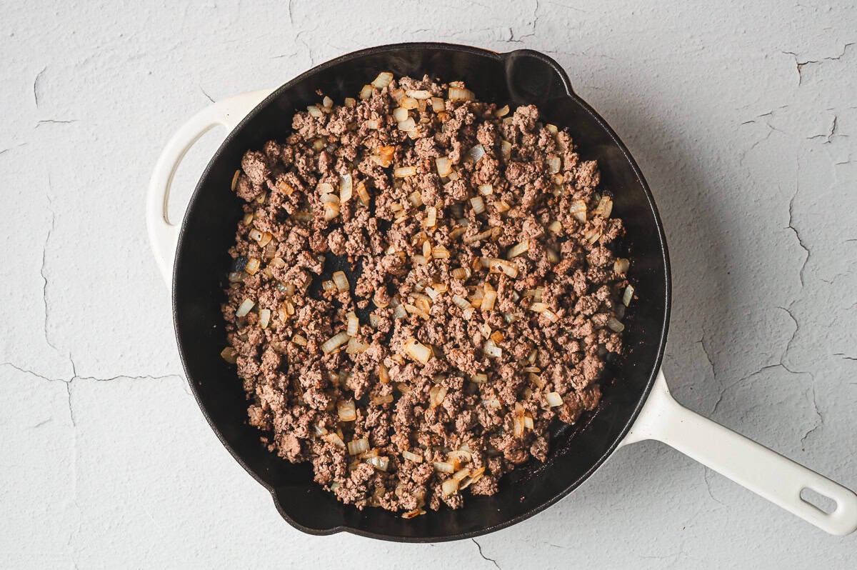 Ground beef and diced onions browning in a black skillet until no pink remains.