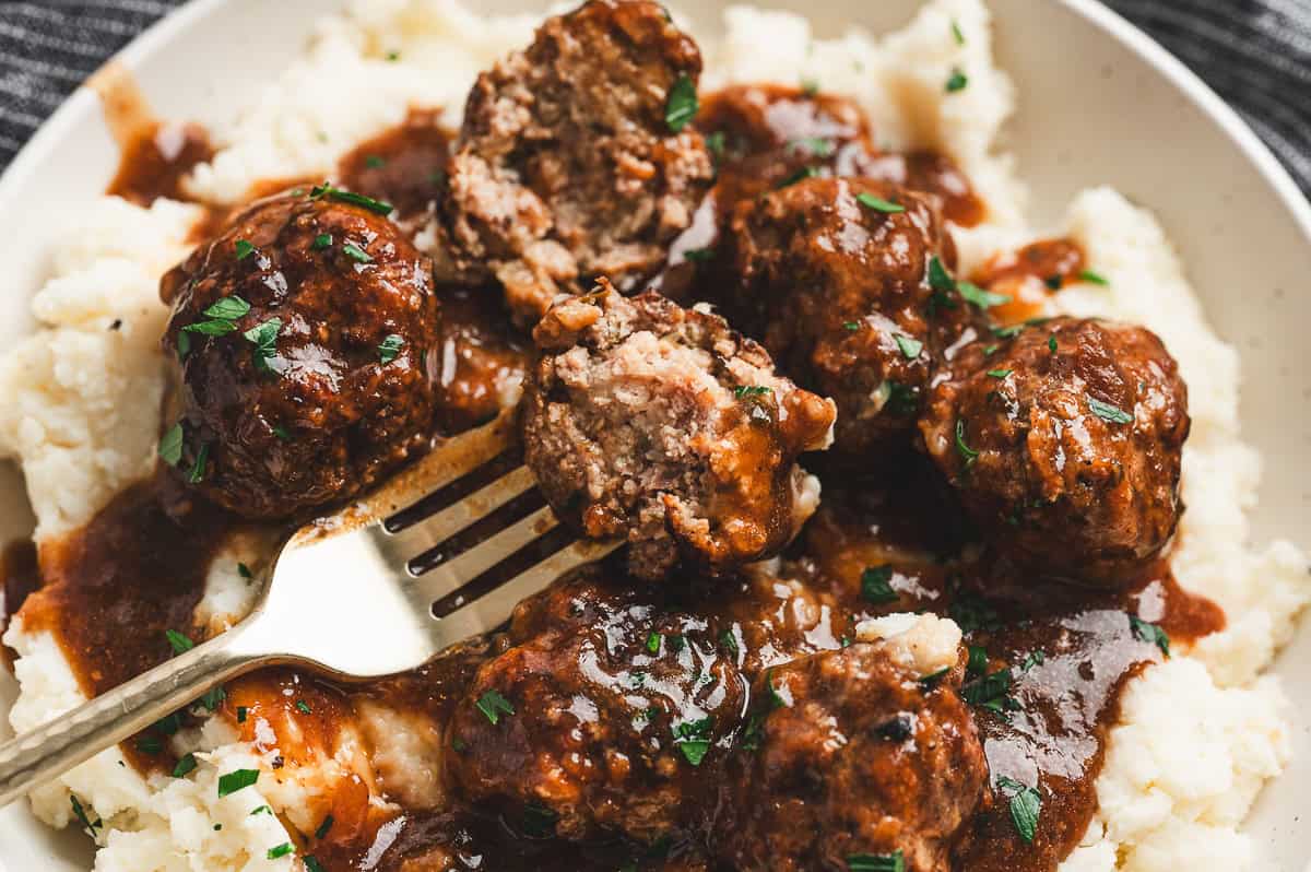 Fork cutting into a Salisbury steak meatball, showing the tender inside, served over mashed potatoes with gravy and parsley.