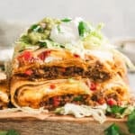 Stacked squares of Crunchwrap Casserole showing beef, cheese, and red peppers, topped with shredded lettuce, sour cream, guacamole, and cilantro.