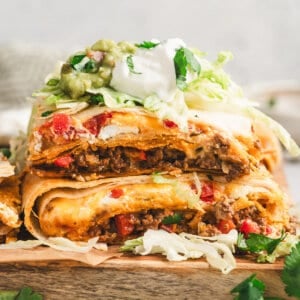 Stacked squares of Crunchwrap Casserole showing beef, cheese, and red peppers, topped with shredded lettuce, sour cream, guacamole, and cilantro.