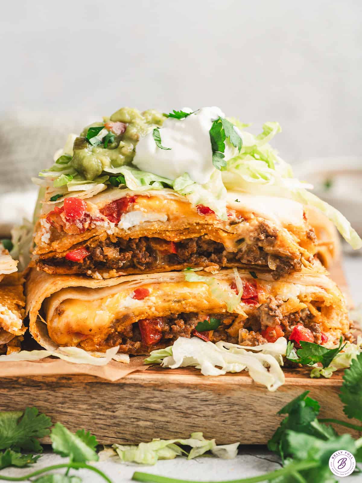 Stacked squares of Crunchwrap Casserole showing beef, cheese, and red peppers, topped with shredded lettuce, sour cream, guacamole, and cilantro.