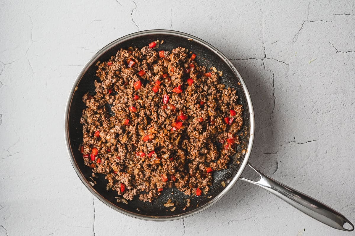 Large skillet filled with browned ground beef, diced onions, and red bell peppers cooked together for the casserole filling.