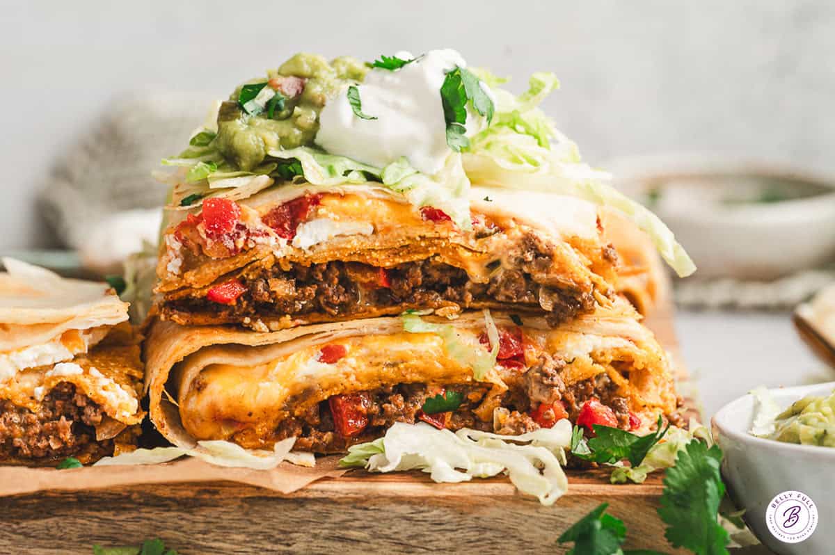 Close-up of stacked slices of Crunchwrap Casserole showing layers of tortillas, beef, cheese, sour cream, and red peppers, topped with lettuce, guacamole, and sour cream.